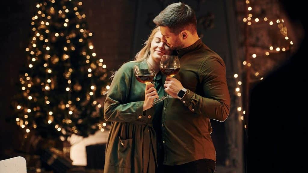 A couple holding wine glasses, embracing near a glowing, out-of-focus Christmas tree.