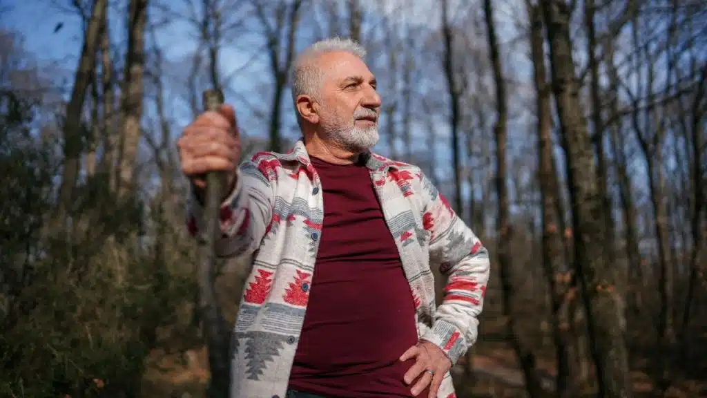 An older man with a white beard holds a stick, standing in a bare forest.