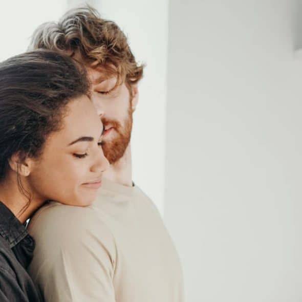 A woman with dark curly hair is hugging a bearded man from behind in a bright indoor setting.