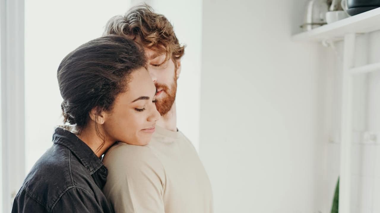 A woman with dark curly hair is hugging a bearded man from behind in a bright indoor setting.