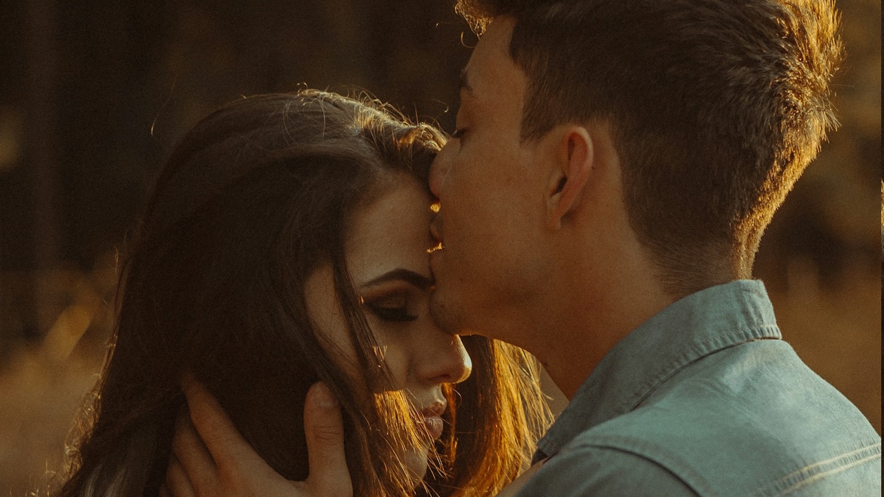 A man kissing a woman's head while standing in nature in the evening.
