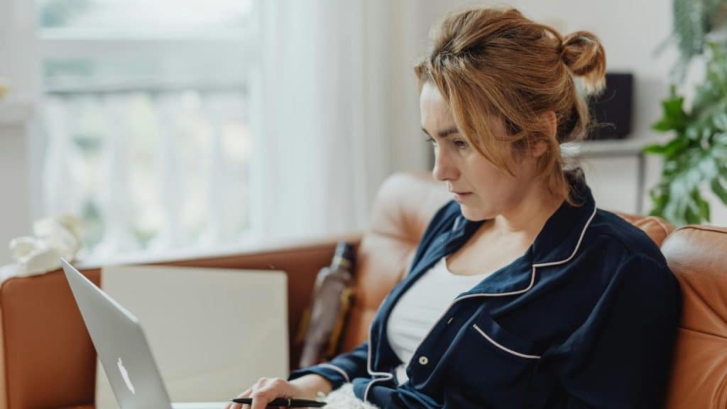 A woman sitting on a couch in pajamas working on a laptop.