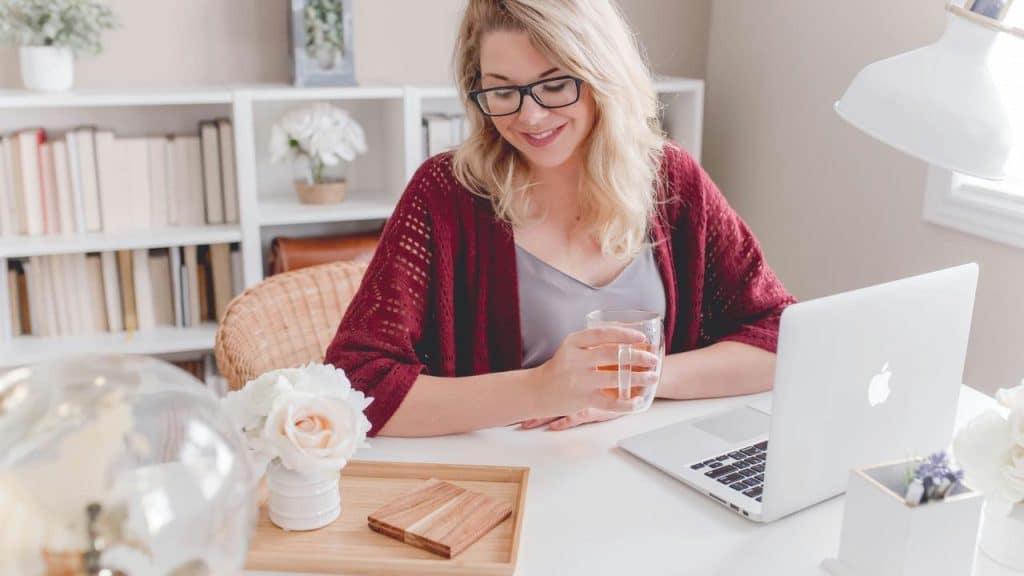 A woman sitting at a desk with a laptop, smiling while holding a glass of tea.