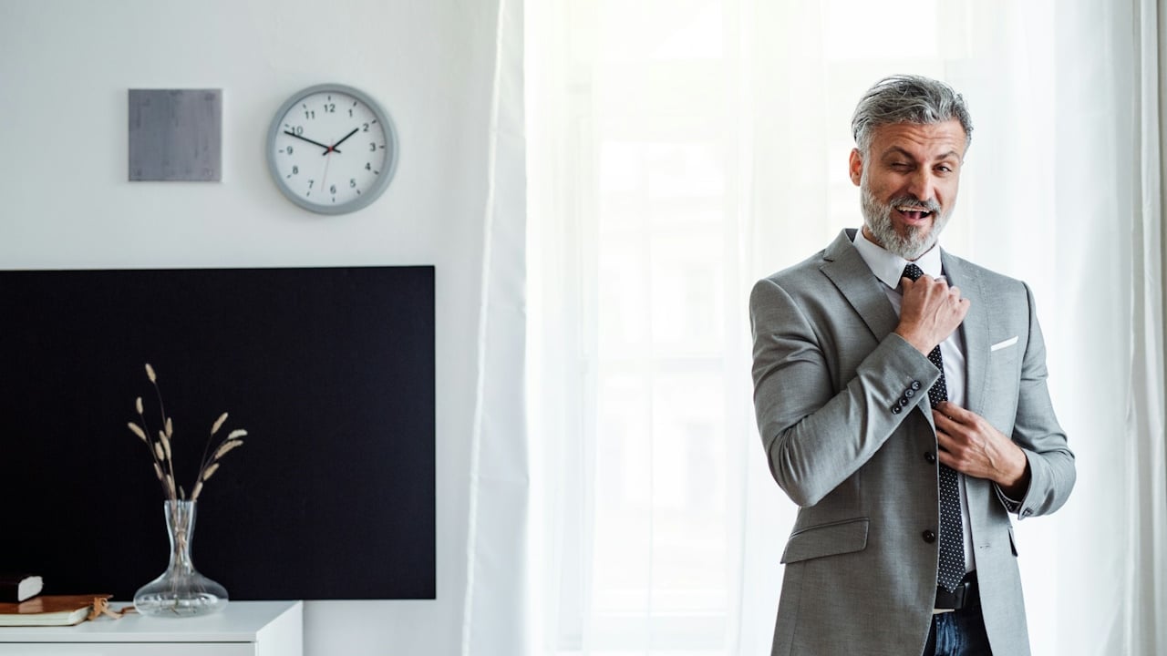 A bearded man in a grey suit is adjusting his tie.
