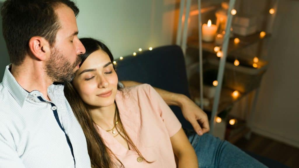 Couple relaxing on a couch, man kisses woman's forehead, string lights in background.