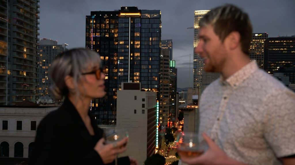 A couple is standing on a balcony with a city skyline at dusk in the background, holding glasses of wine and talking.