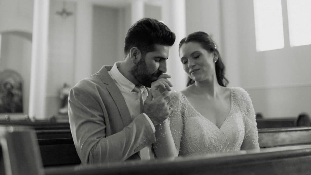 A black and white photo of a groom holding the bride's hands as they sit together in a church pew.