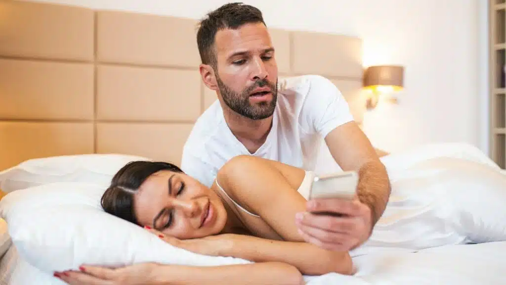 A man leans over a woman in bed, holding a phone and looking at the screen.
