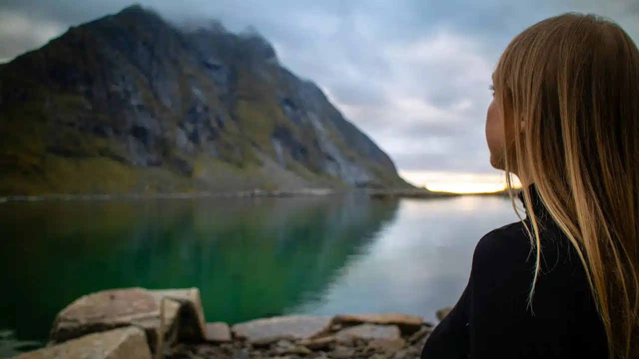 A woman in a black jacket is sitting on a rock near a lake during the daytime.