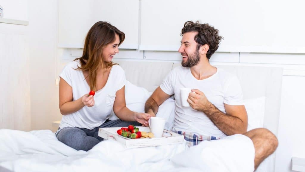 Man and woman smiling and laughing while eating a tray of breakfast in bed.