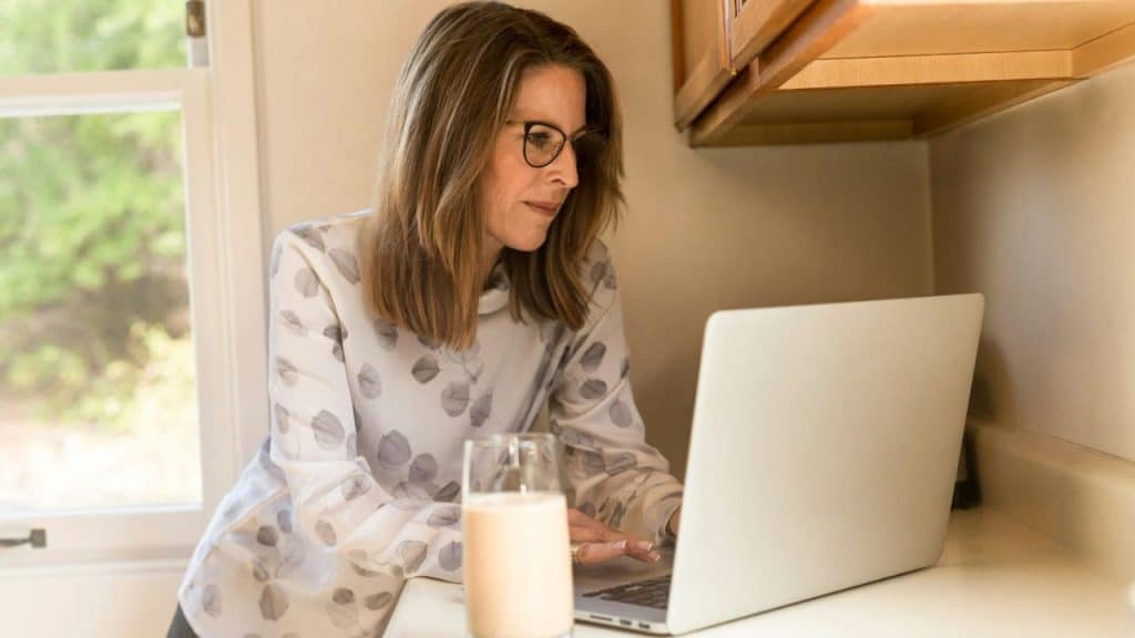 A woman wearing glasses works on a laptop in a kitchen, a glass of liquid nearby.