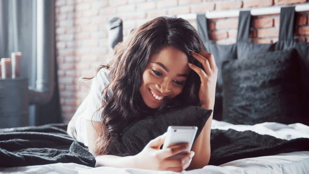Woman lying on her stomach in bed, smiling at her phone.