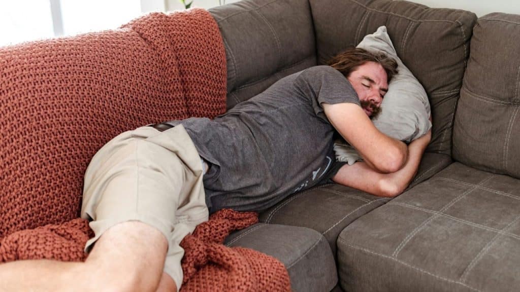 A man is asleep, lying on his side on a large sectional sofa.