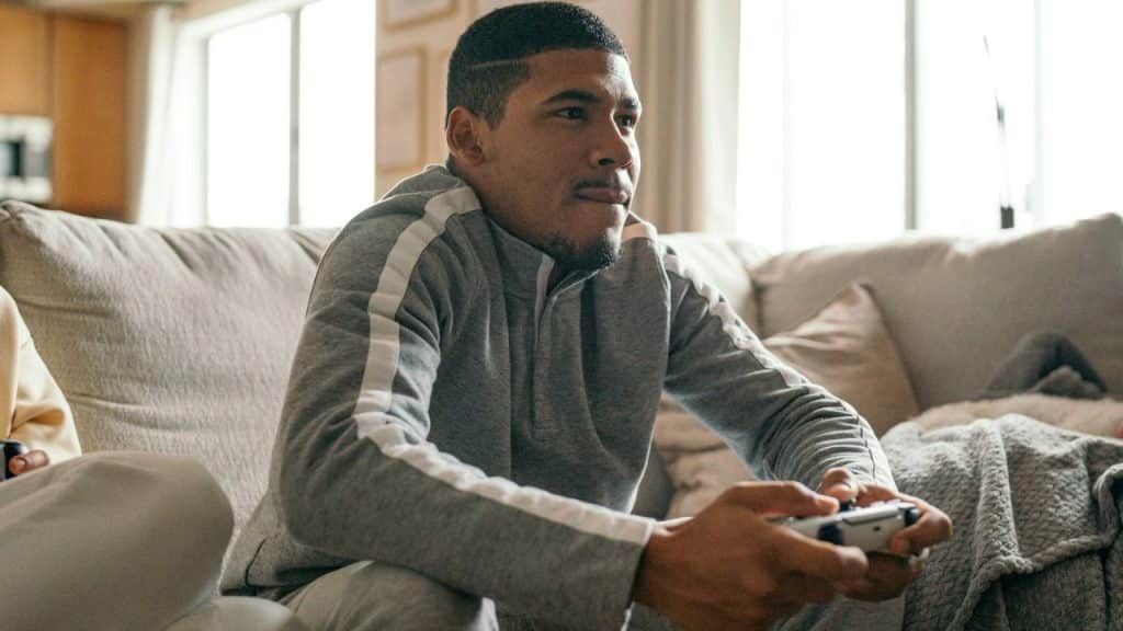 A man in a gray hooded sweatshirt sits on a sofa and plays a video game intensely.