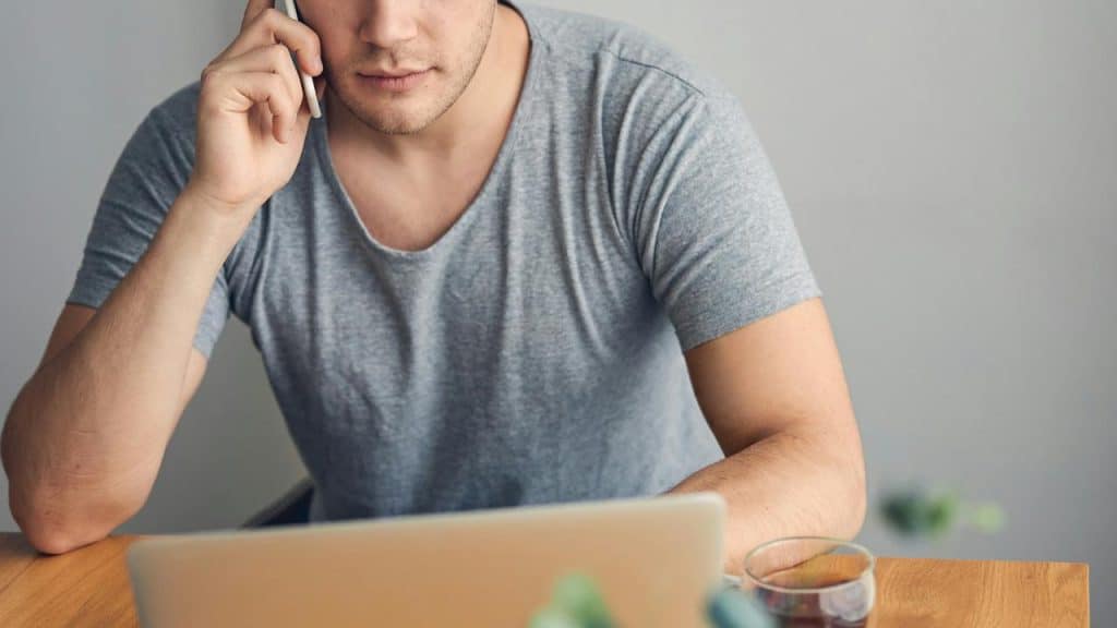 A man in a gray shirt talking on the phone while using a laptop.