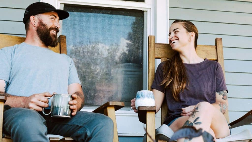 Man and woman sitting in rocking chairs on a porch, laughing and holding coffee mugs.