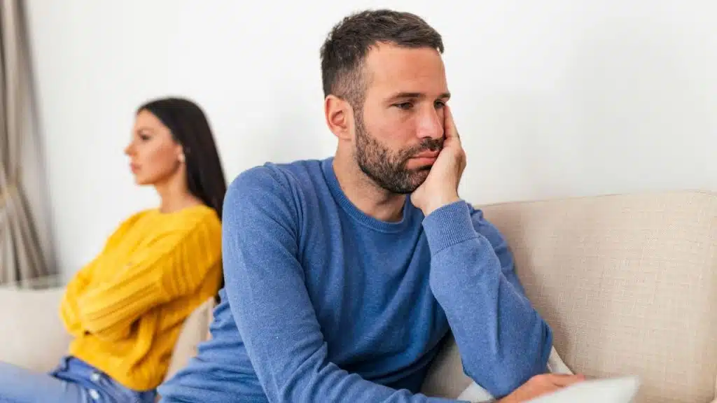 A sad man sits with his hand on his face, back to a woman on a couch.