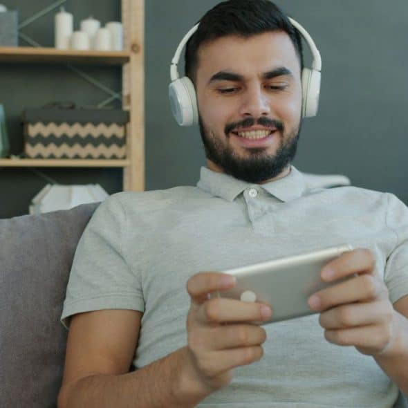 Man with headphones smiling while looking at a phone on a couch.