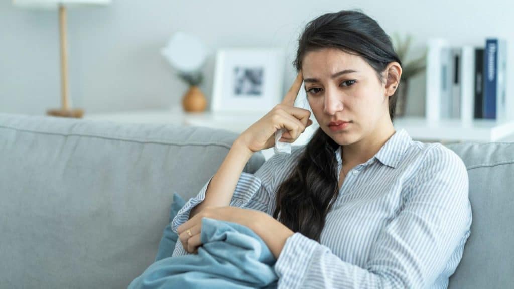 Woman sitting on a couch, holding a blanket and touching her forehead thoughtfully.