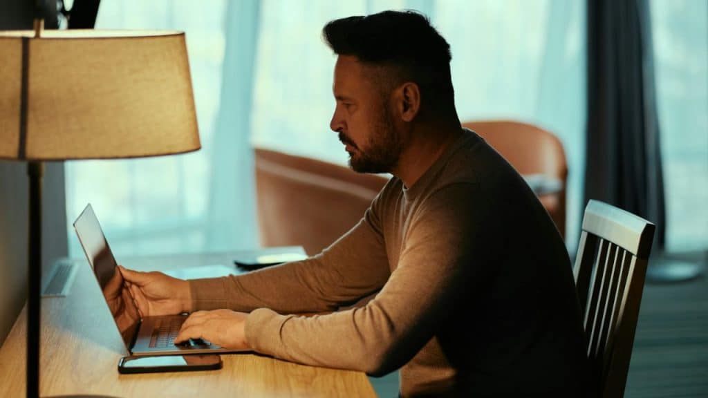Man sitting at a desk with a lamp, looking intently at a laptop keyboard.