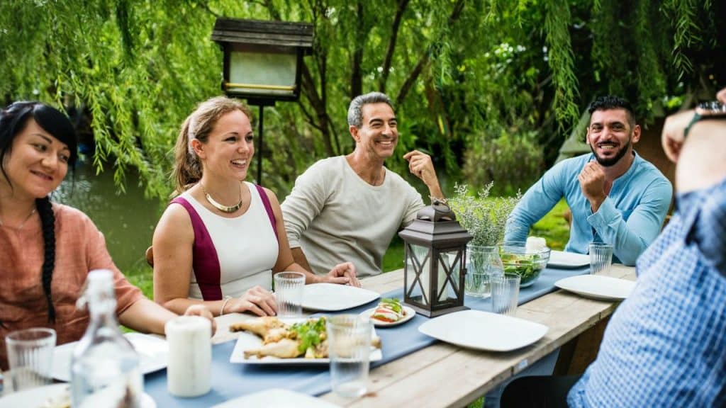 Four smiling adults sitting at a wooden outdoor dining table with plates and food.