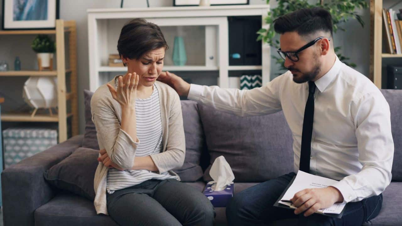 A man comforts a crying woman on a couch, offering a supportive pat on the shoulder.