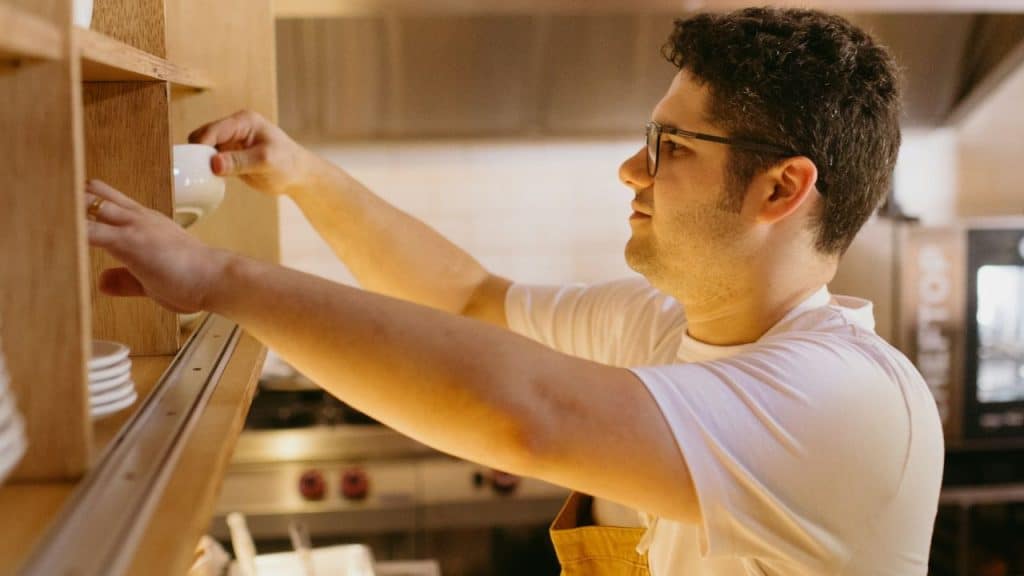 Man in glasses and apron placing a small white cup on a wooden shelf.