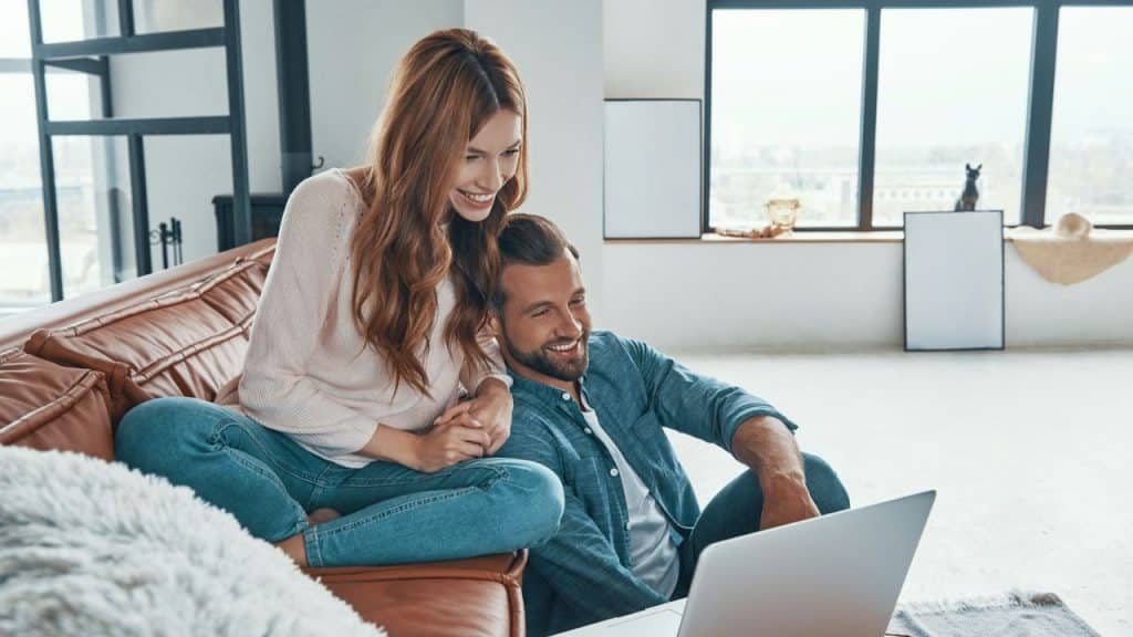 Smiling woman sitting on a couch and man sitting on the floor using a laptop.