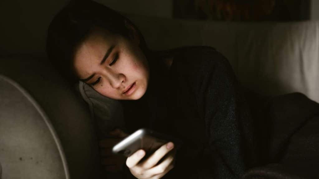 A woman lying on a couch while looking at her phone in dim light.