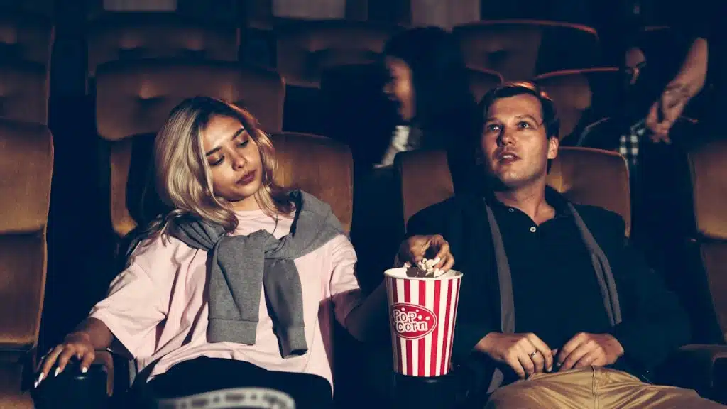Man eating popcorn in a dark movie theater while a woman next to him appears to be asleep.
