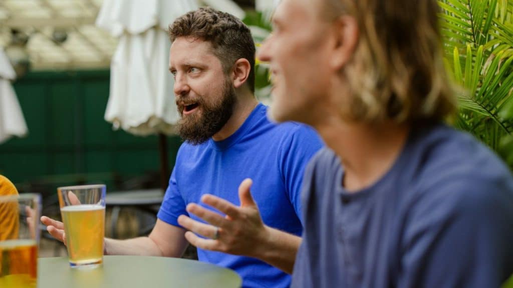 Two men talking at an outdoor table with drinks, one wearing a blue shirt.