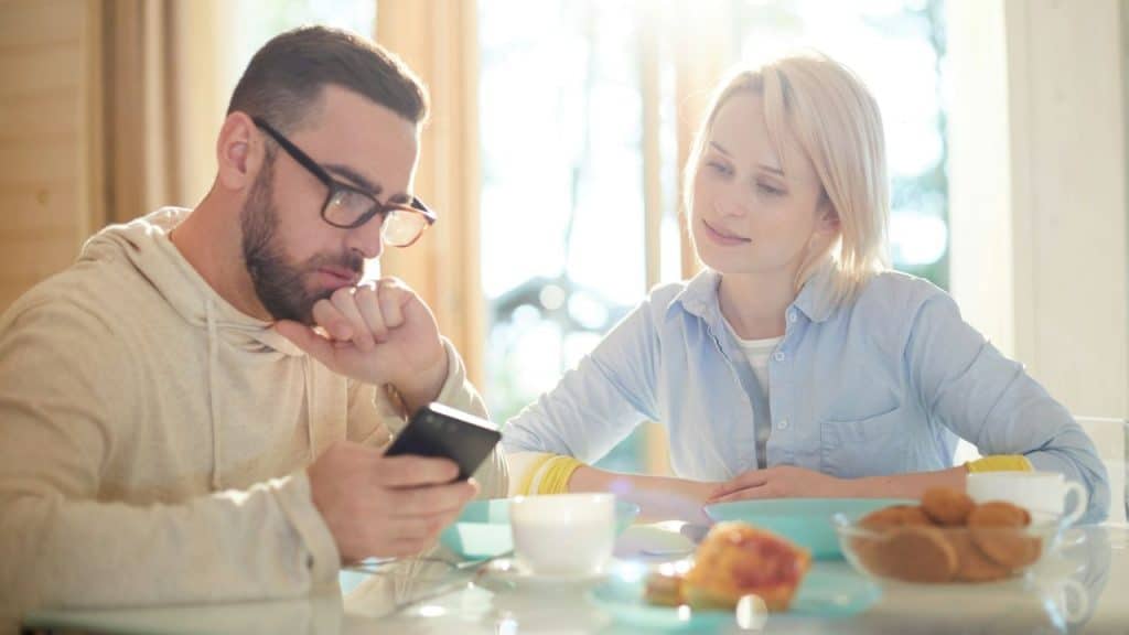 A man in glasses looks at a phone while a woman watches him across a bright table with food.