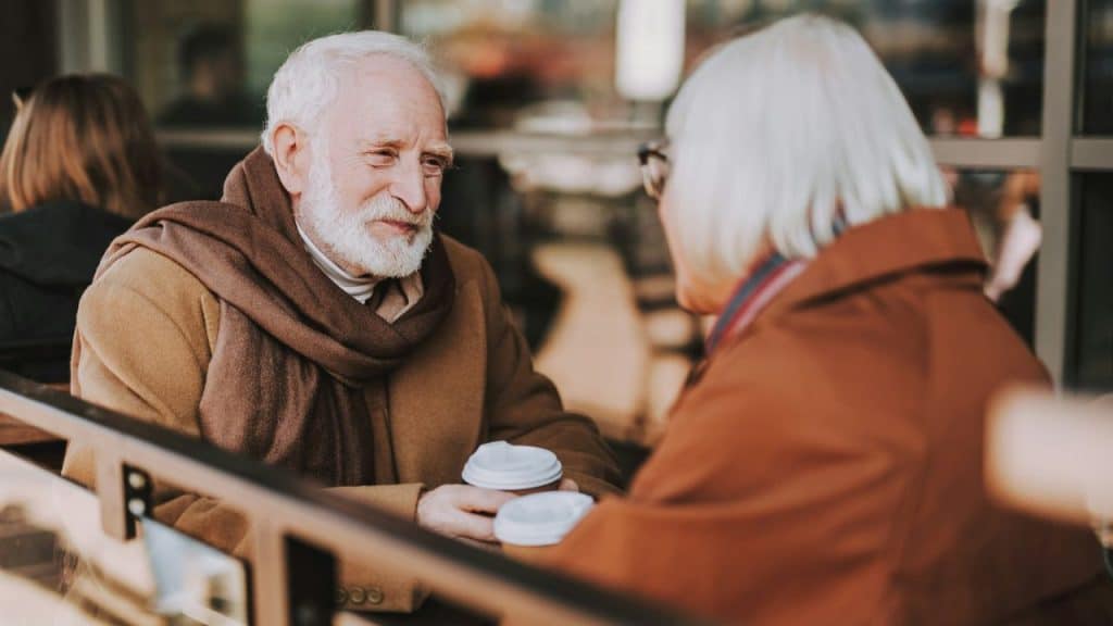 Elderly man wearing a coat and scarf talks with a woman while holding coffee cups.