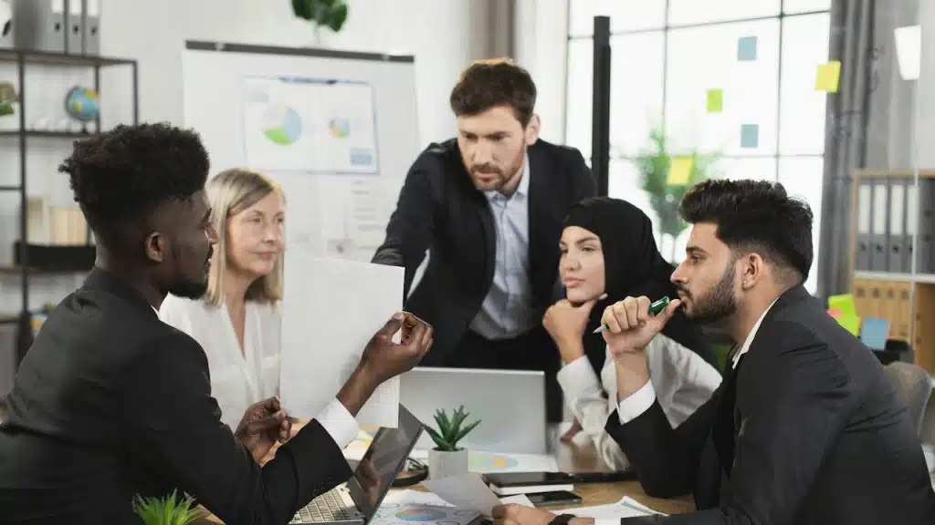 Five business professionals, including a woman in a hijab, discussing documents around a table in an office setting.