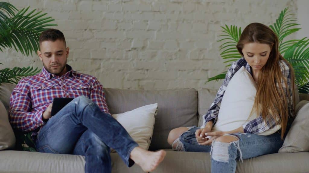 A couple sits separately on a couch, the man on a tablet, the woman looking down sadly.