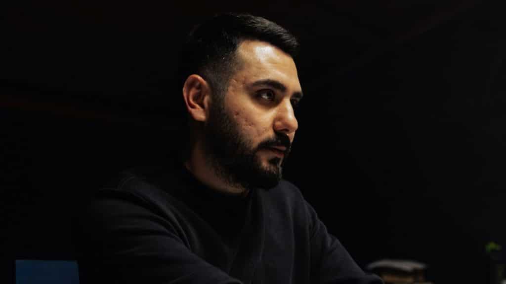 A man with dark hair and a beard sits in a dimly lit room, looking away.