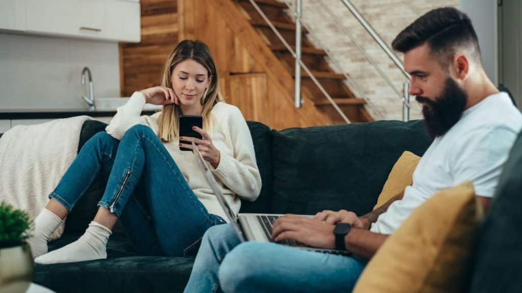 A man sits with a laptop while a woman uses her phone on a couch.