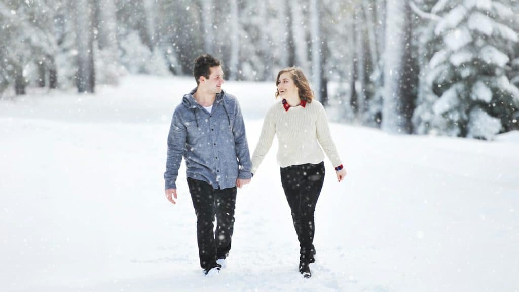 A smiling young couple holding hands while walking through a snowy forest.