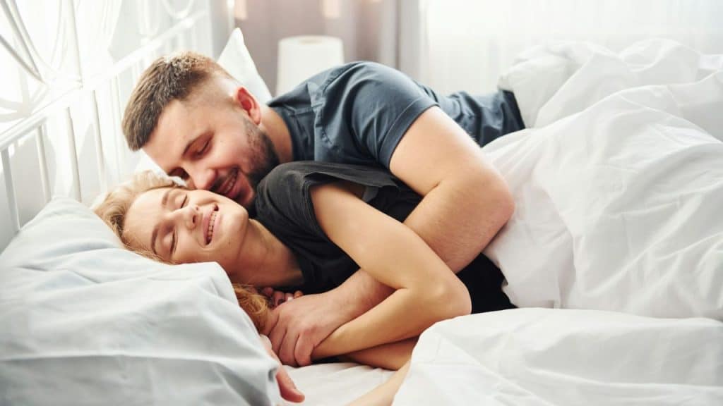 A smiling couple cuddles in bed, illuminated by sunlight.