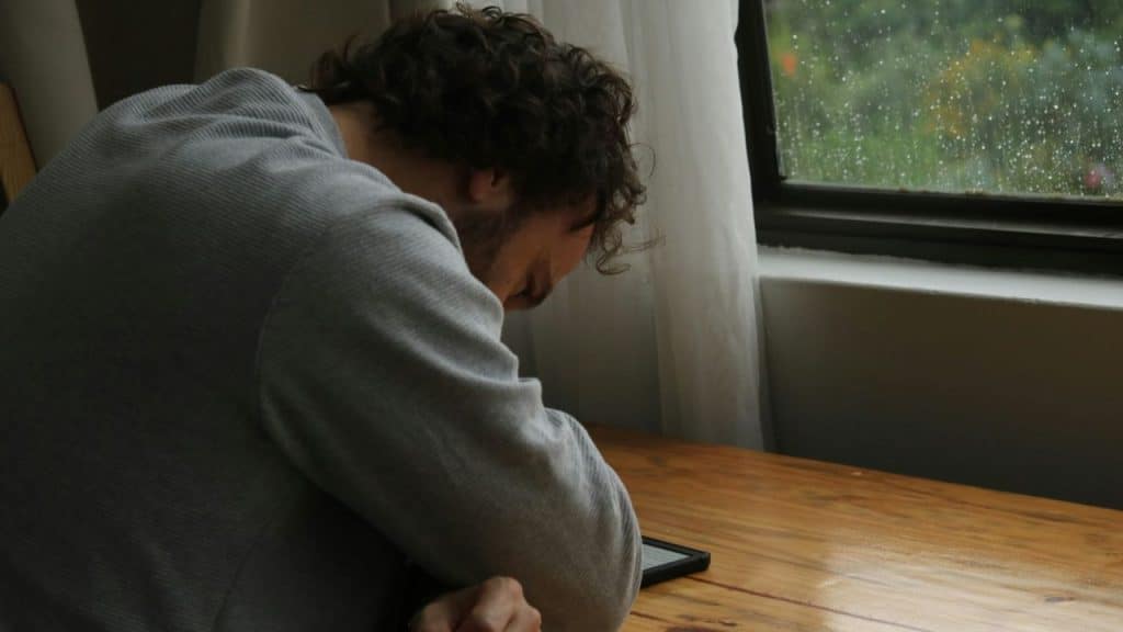 Man slumped over a wooden table, looking at a small electronic device.