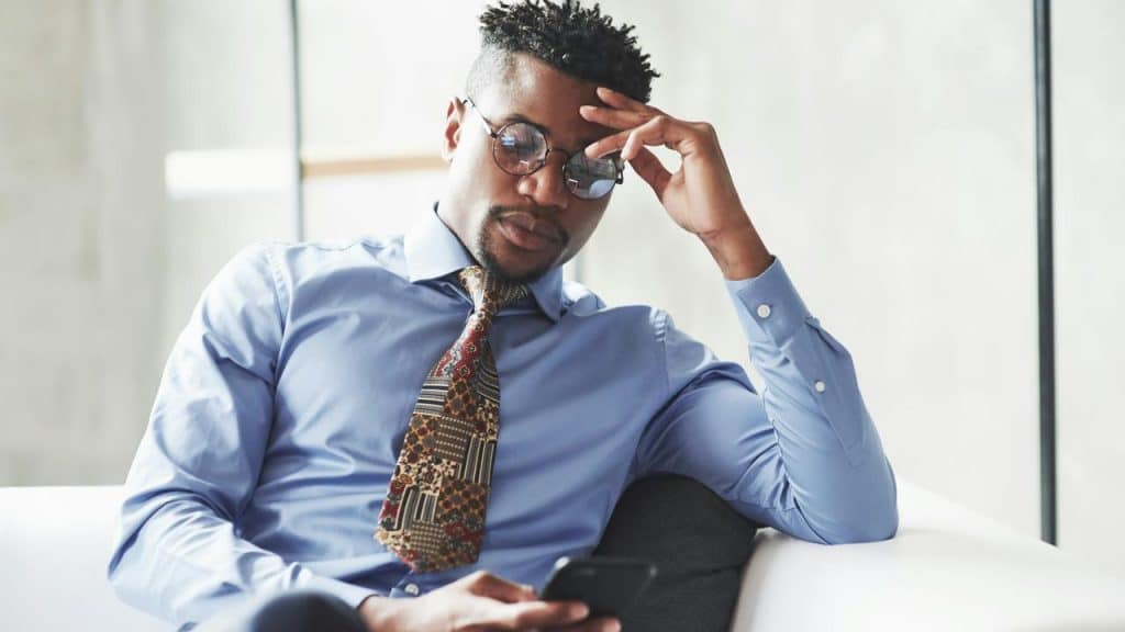 A man in a blue shirt and patterned tie sitting on a couch looking at his phone thoughtfully.