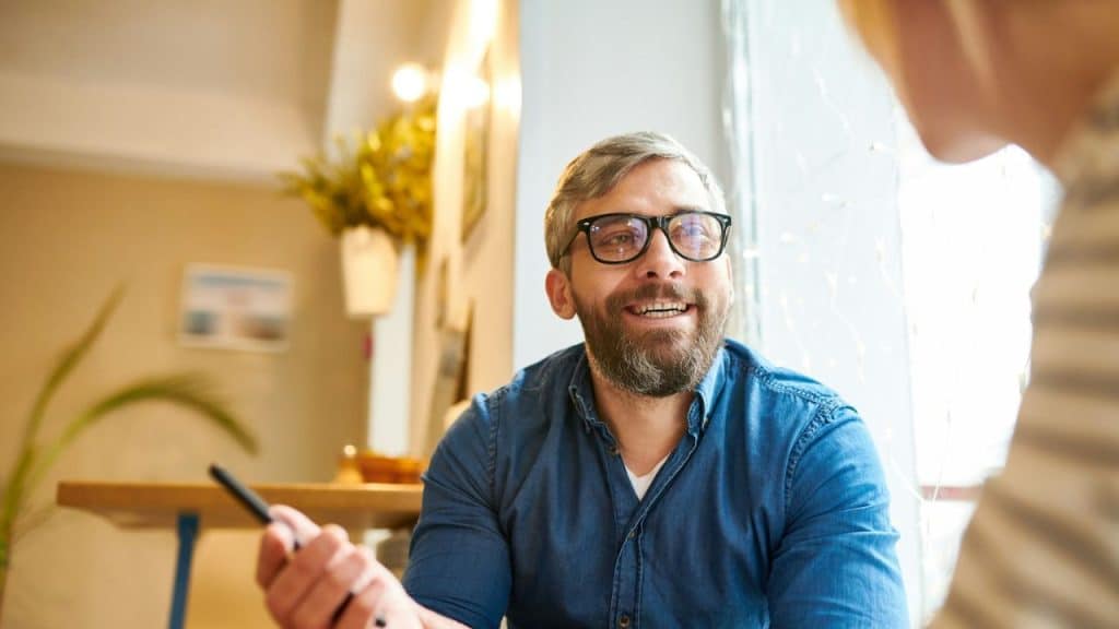 Man with glasses and beard smiling while holding a pen in a sunlit cafe.