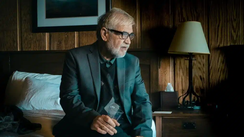 Elderly man in a suit jacket and glasses sitting on a bed holding a glass.
