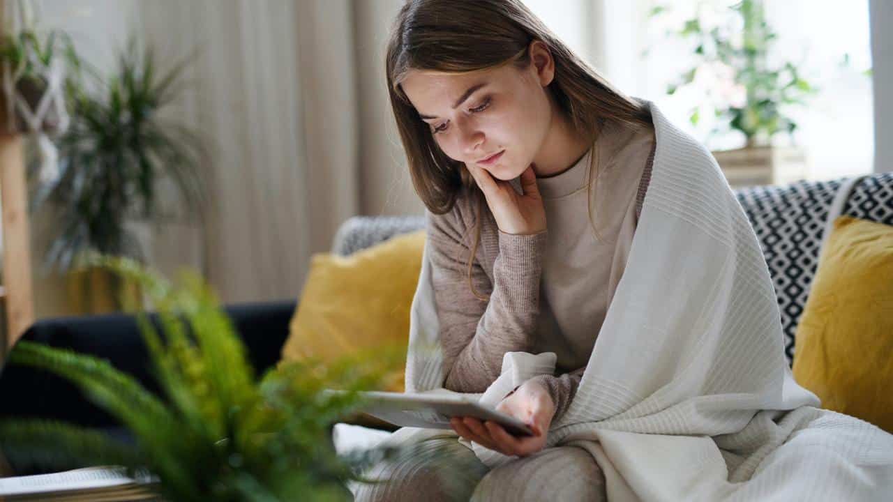 A young woman sitting on a couch wrapped in a blanket, reading a book.