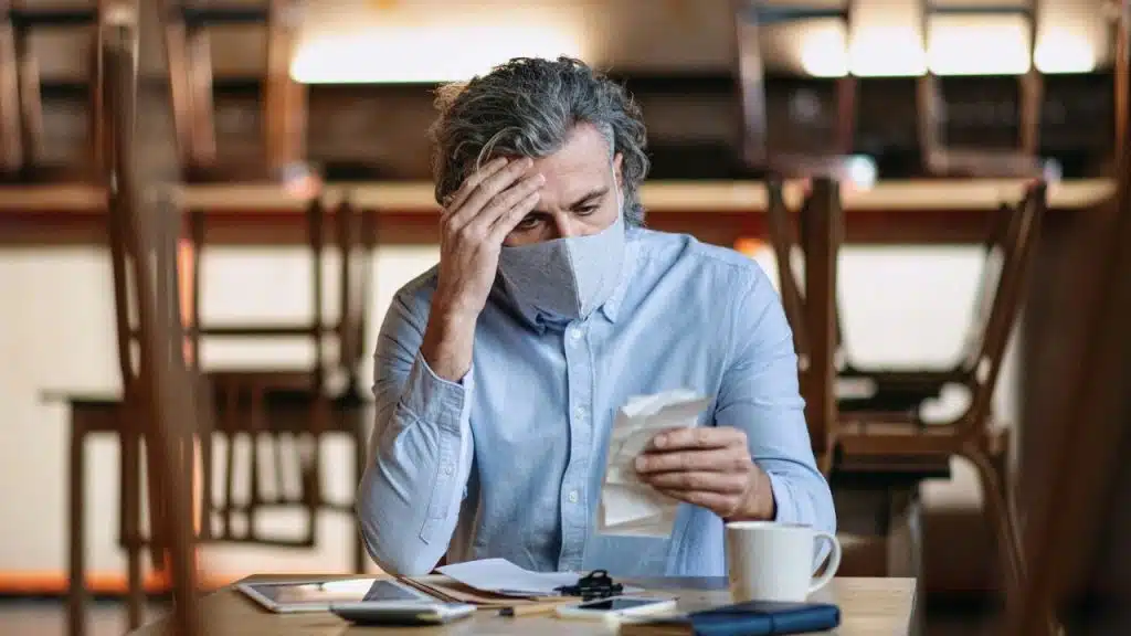 Man with a mask sits at a table, holding a crumpled paper with his hand on his head.