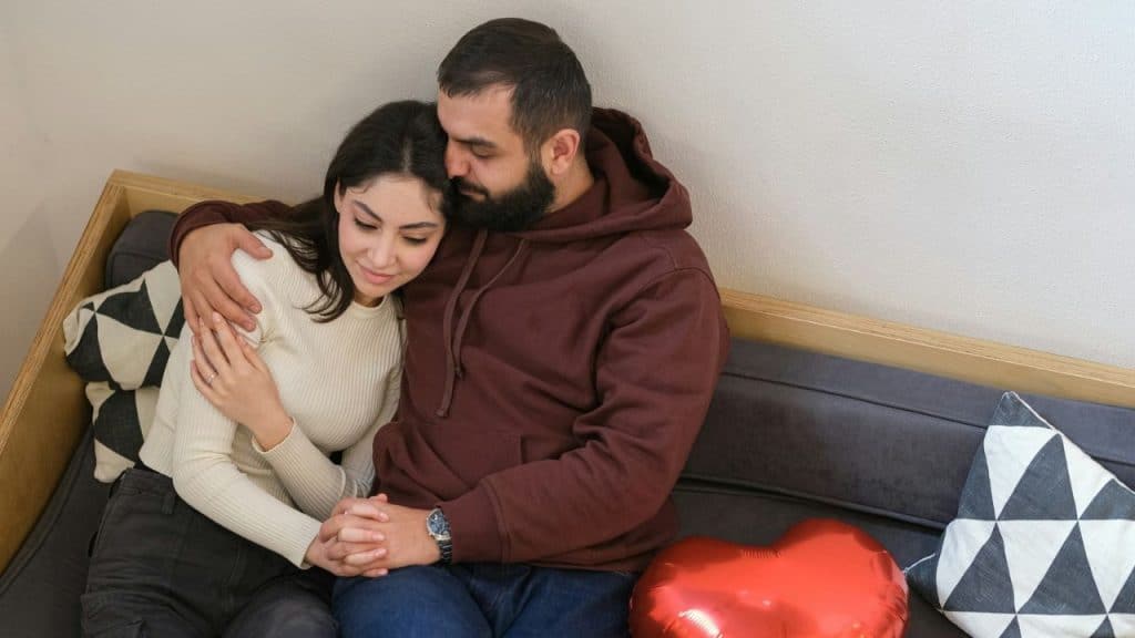 Man with a beard hugging a woman on a couch next to a heart-shaped balloon.