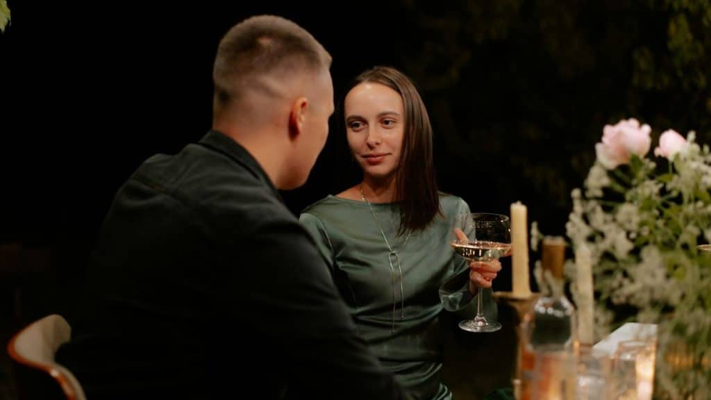 A woman in a silk dress holding a wine glass, smiling at a man across a table.
