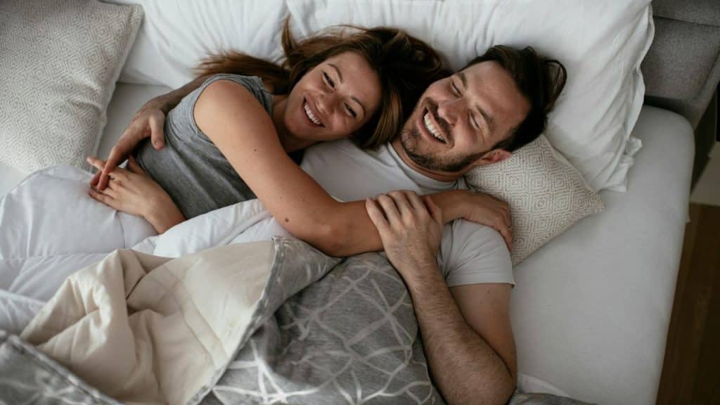A man and woman lie in bed together under a blanket, smiling and hugging.