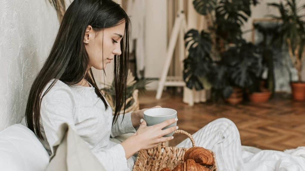 A woman sitting on a bed holding a cup with a basket of croissants.