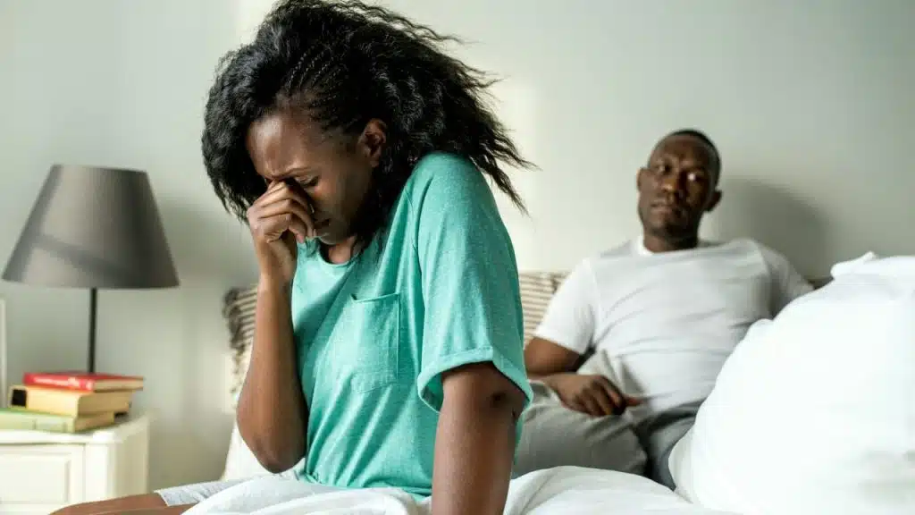 A woman sits on a bed crying into her hand while a man watches from behind.